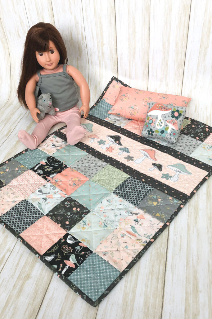 Patchwork doll quilt by top US sewing blog Ameroonie Designs. Image of doll with quilt and pillows.