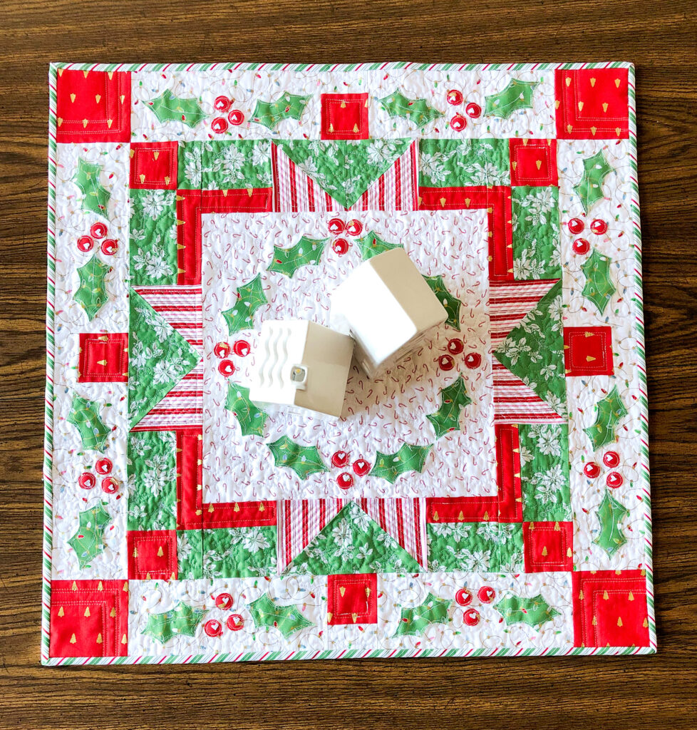 Delightful rainbow mini quilt by top US sewing blog Ameroonie Designs. Image of table topper with holly applique.