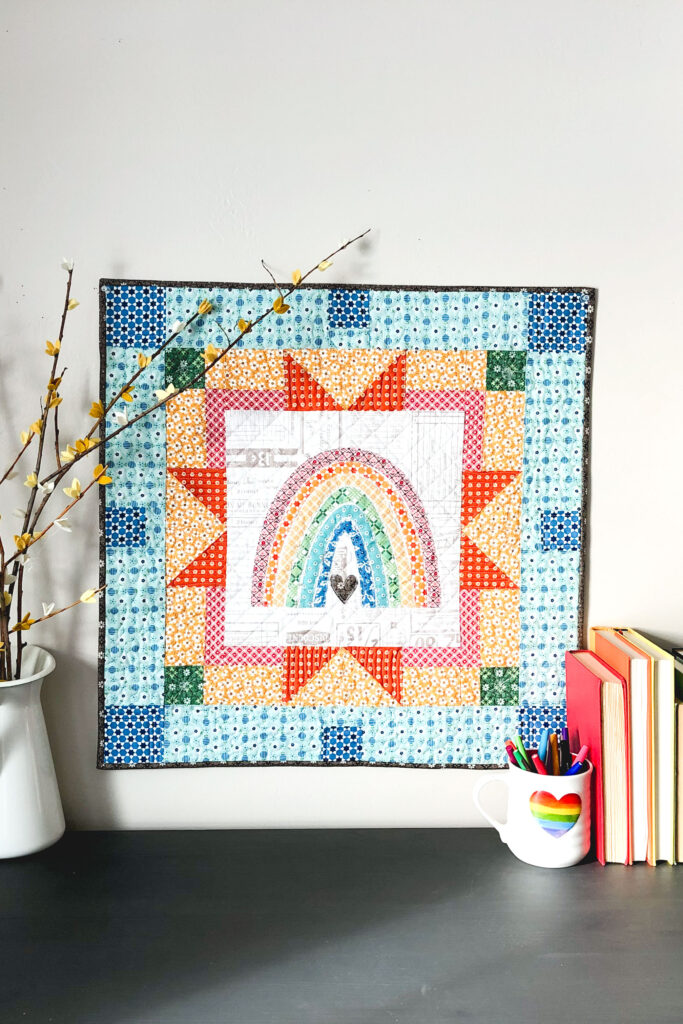 Delightful rainbow mini quilt by top US sewing blog Ameroonie Designs. Image of rainbow mini quilt with books and branches.