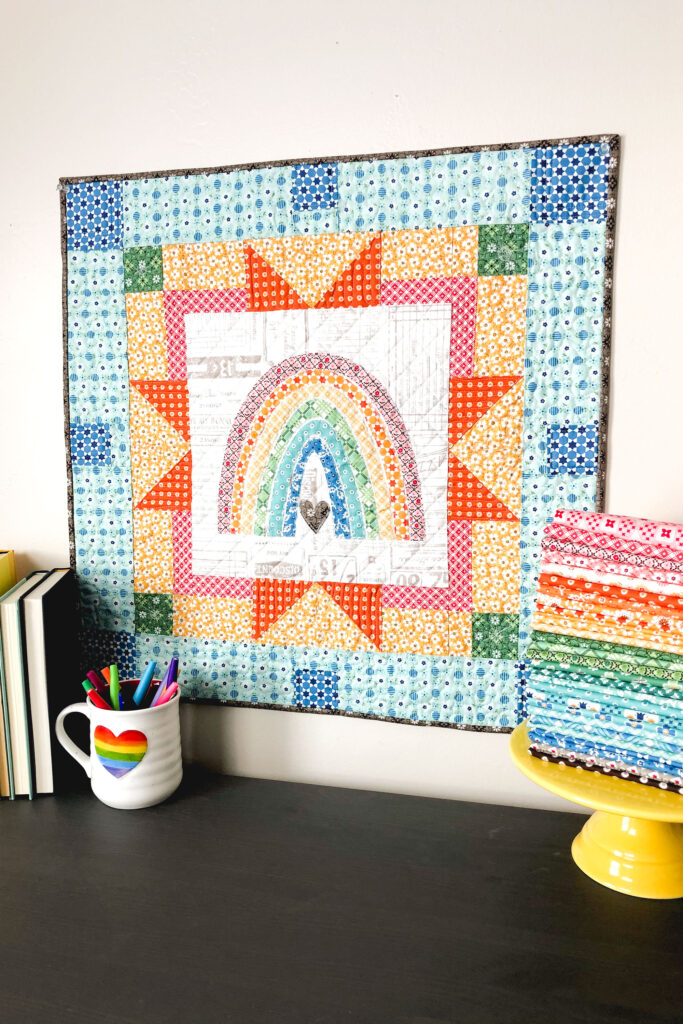 Delightful rainbow mini quilt by top US sewing blog Ameroonie Designs. Image of mini quilt with rainbow applique center.
