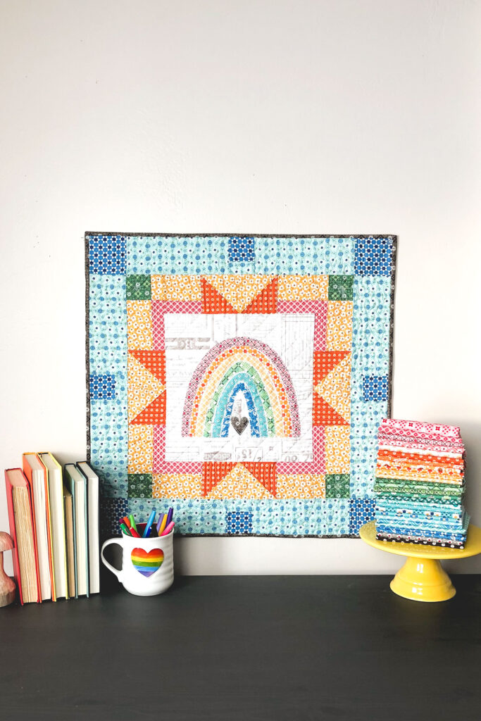 Delightful rainbow mini quilt by top US sewing blog Ameroonie Designs. Image of rainbow applique mini quilt with fabric and books.