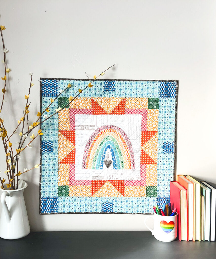 Delightful rainbow mini quilt by top US sewing blog Ameroonie Designs. Image of mini quilt with books and branches.