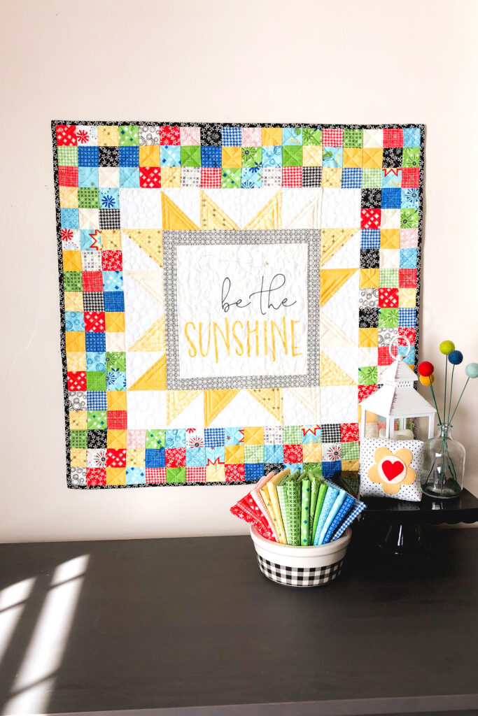Sew a stunning mini quilt with a fabric panel by top US sewing blog Ameroonie Designs. Image of mini quilt on wall with sunlight.