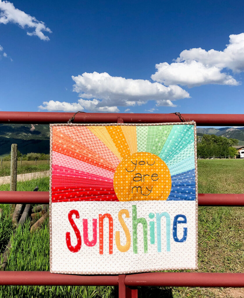 Rainbow Sunshine mini quilt sew along with top US sewing blog Ameroonie Designs. Image of mini quilt on barn fence and sky.