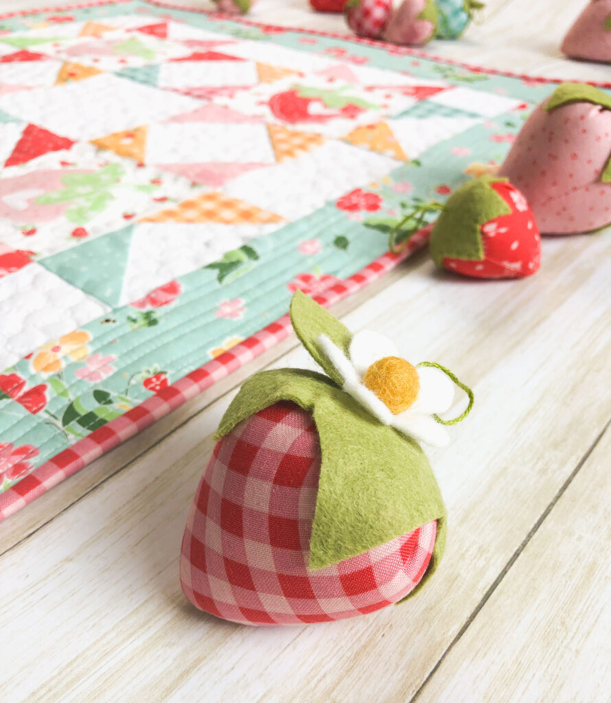 Strawberry Springtime mini quilt free pattern by top US sewing blog Ameroonie Designs. Image of stuffed fabric strawberry with mini quilt and more strawberries in background.