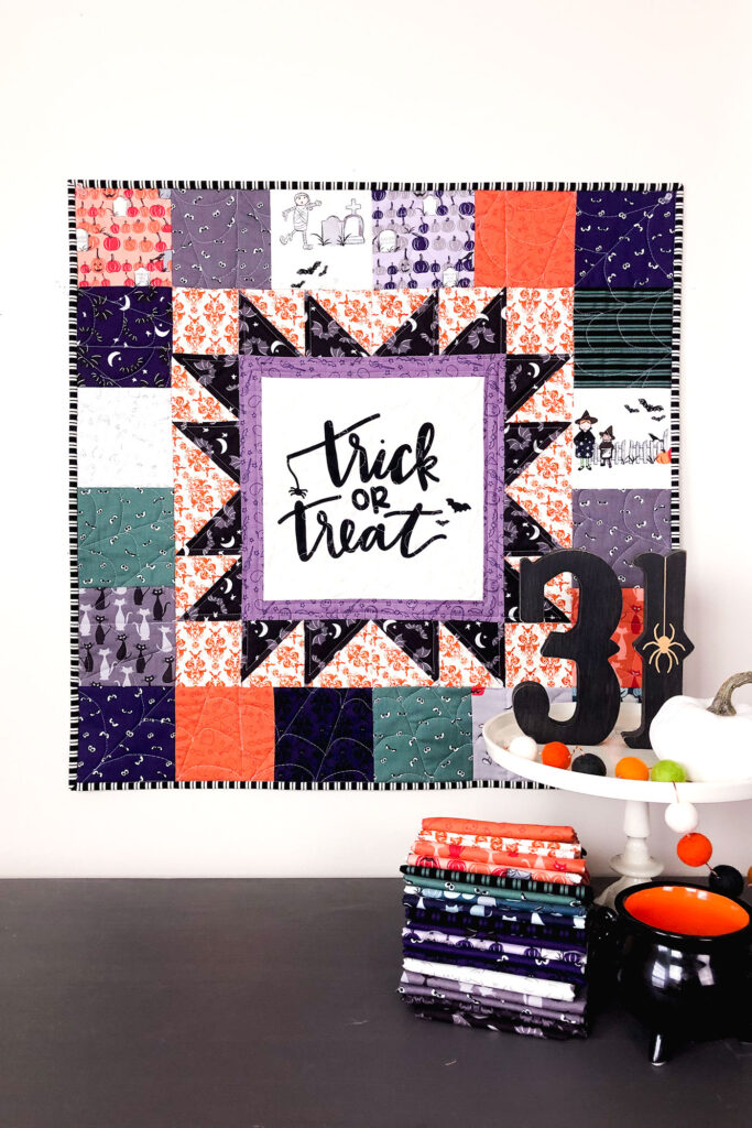 Turn a fabric panel into a stunning mini quilt from Top US sewing blog Ameroonie Designs. Image of mini quilt on wall with fabric and Halloween decorations.