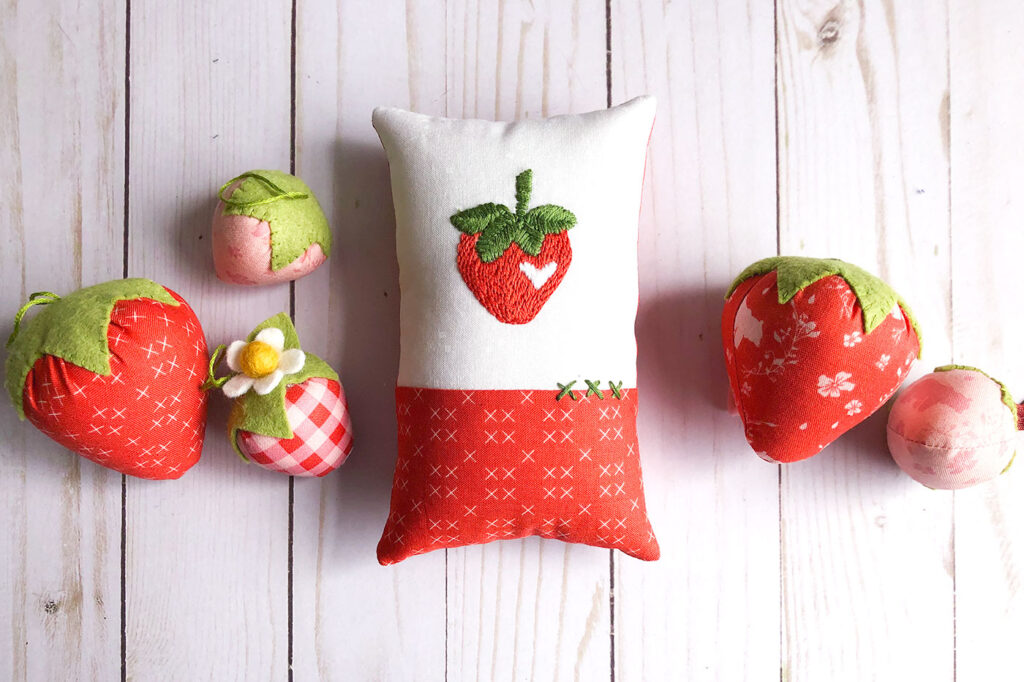 How to sew an easy embroidered strawberry mini pillow with top US sewing blog Ameroonie Designs. Image of mini pillow surrounded by fabric strawberries.