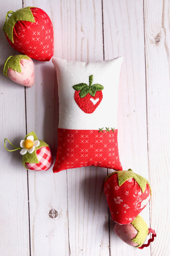 How to sew an easy embroidered strawberry mini pillow with top US sewing blog Ameroonie Designs. Image of mini pillow surrounded by fabric strawberries.