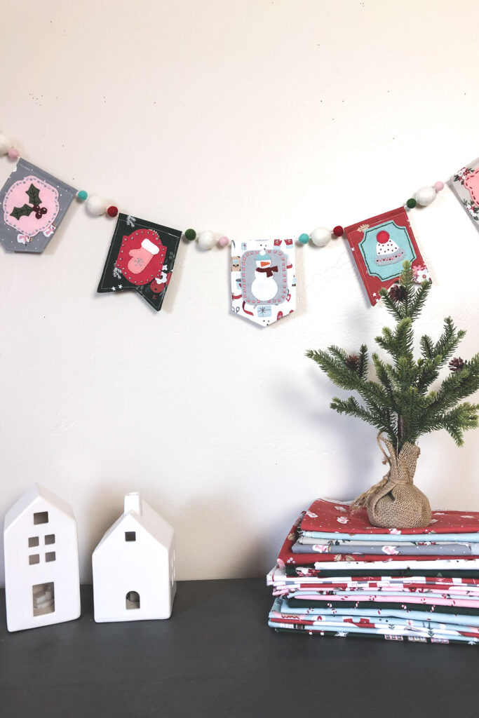 Gorgeous winter mini banner sneak peek with top US sewing blog Ameroonie Designs. Image of mini banner strung with pom poms, houses and mini pine tree.