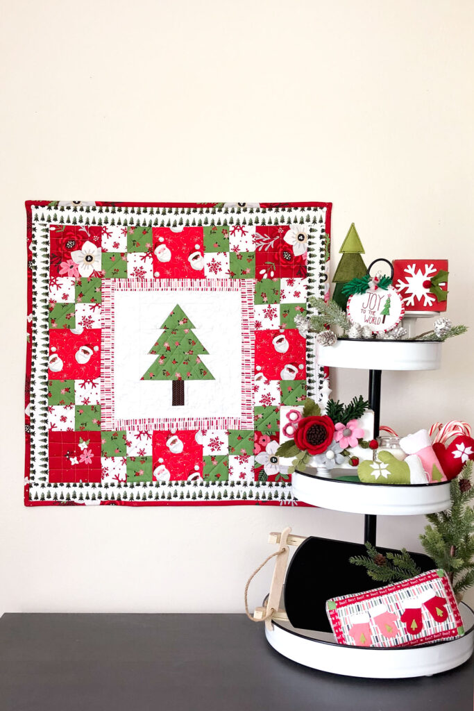 How to sew a delightful holiday mini quilt by top US sewing blog Ameroonie Designs. Image of holiday scene with mini quilt and decorated tiered tray.