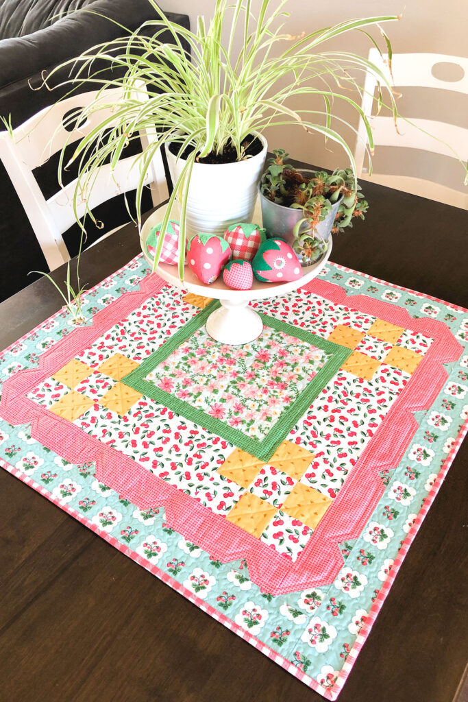 Stunning Scalloped Table Topper by top US sewing blog Ameroonie Designs. Image of table topper on table with cake plate, plants and stuffed fabric strawberries.