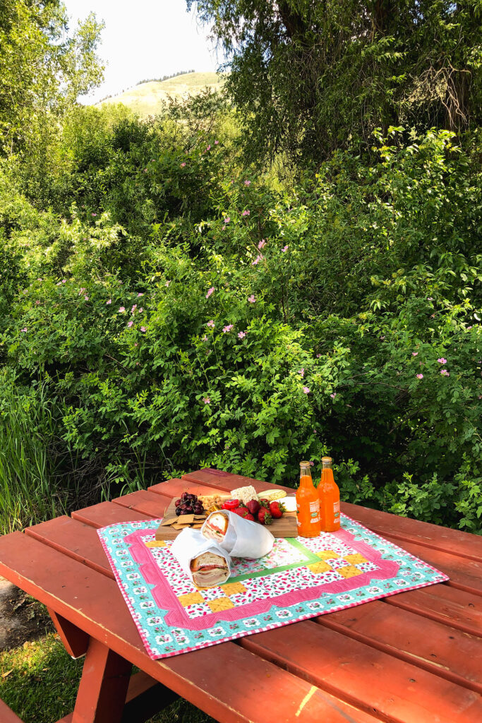 Stunning Scalloped Table Topper by top US sewing blog Ameroonie Designs. Image of table topper with picnic on table with mountain and greenery in background.