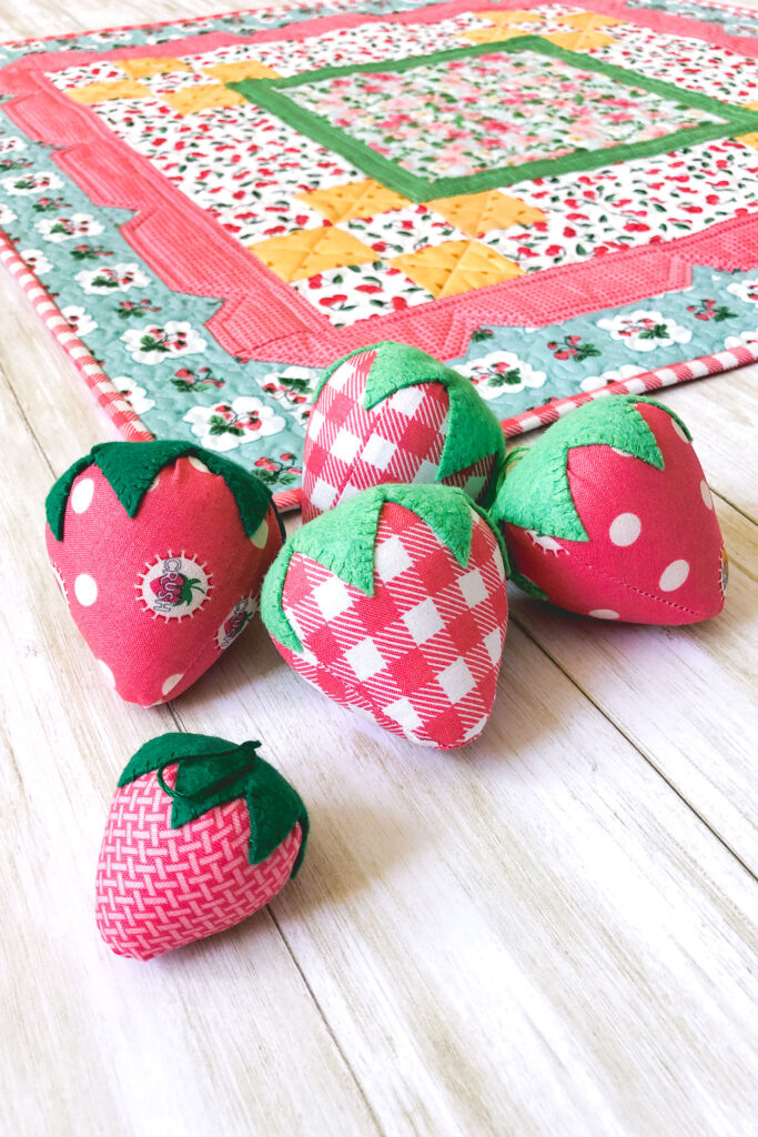 Stunning Scalloped Table Topper by top US sewing blog Ameroonie Designs. Image of fabric strawberries in front of table topper.