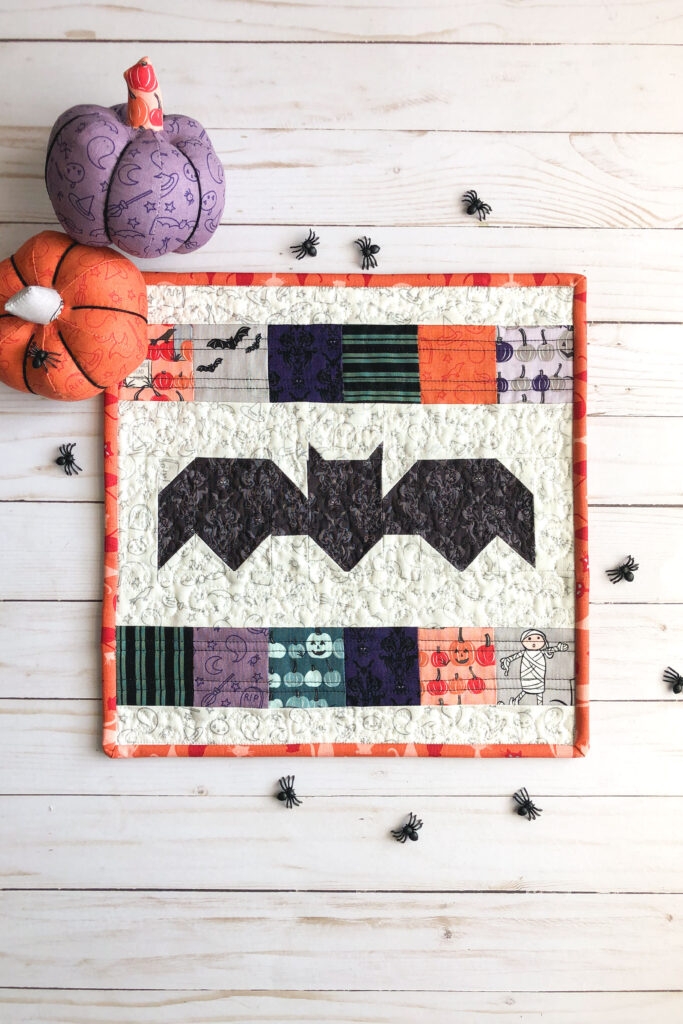 New Halloween Sewing patterns by top US sewing blog Ameroonie Designs. Image of bat mini quilt with fabric pumpkins and plastic spiders.