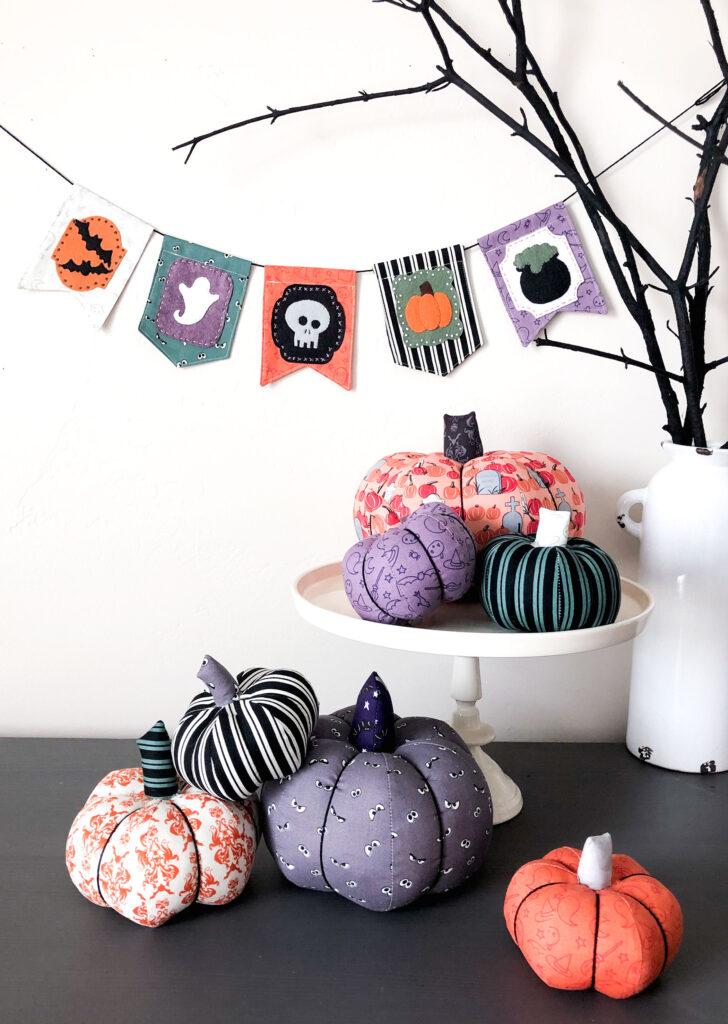 New Halloween Sewing patterns by top US sewing blog Ameroonie Designs. Image of fabric pumpkins in front of black branches with a Halloween banner hanging in the background.